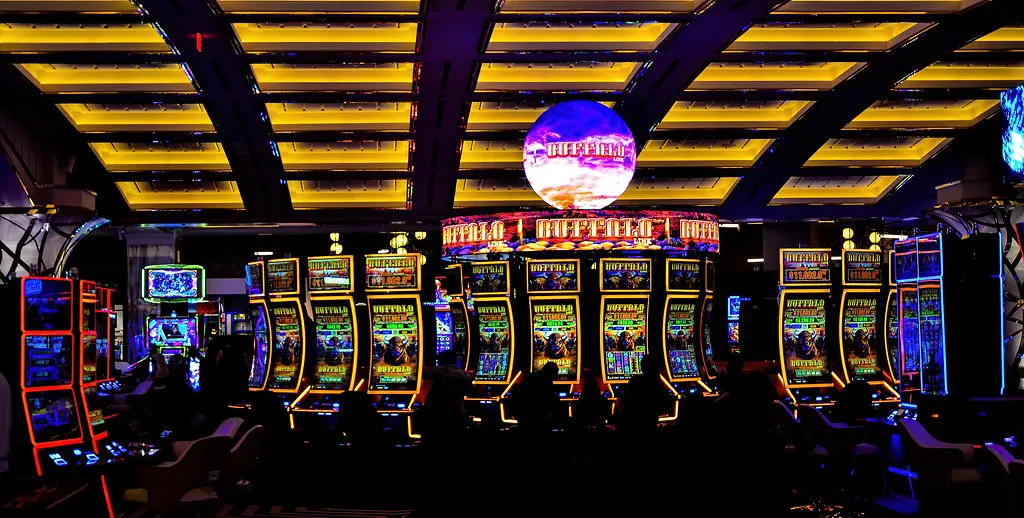 Rows of illuminated slot machines on a casino floor under a yellow geometric ceiling.